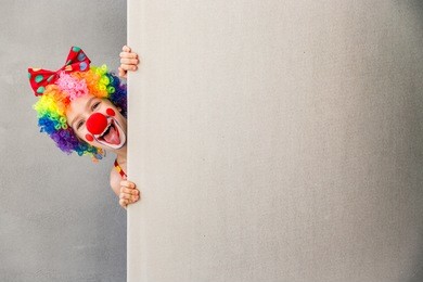 let's celebrate! funny kid clown hiding behind cardboard blank. child holding banner with copy space for your text. 1 april fool's day concept