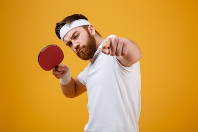 photo of handsome serious young sportsman dressed in white t-shirt standing isolated over yellow background holding racket for table tennis. looking at camera while pointing.