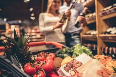 at the supermarket. shopping cart full of goods, in the background young couple choosing wine