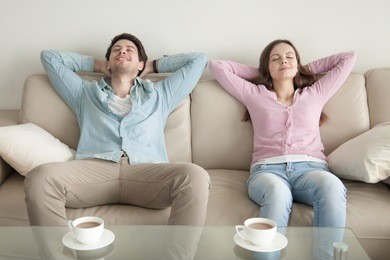 happy young couple enjoying sitting on sofa at home, resting hands behind the head, relaxing with eyes closed, cup of coffee in the morning, dreaming of vacation, listening to music, leisure lazy day