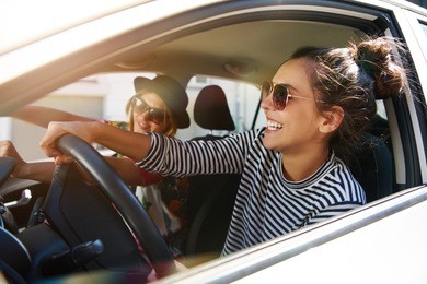 two fun young women in sunglasses driving in a car in town laughing and smiling as they socialise together, view through open side window