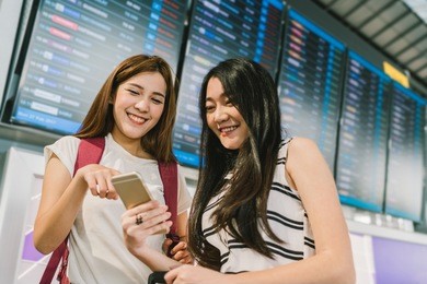 two asian girls using smartphone together at flight information board in airport. online check-in, timetable application, or holiday travel concept