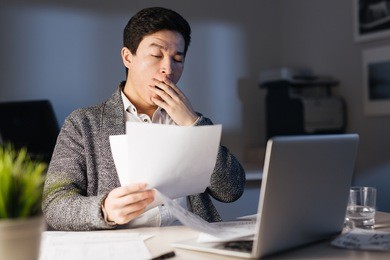 portrait of sleepy asian man yawning while working overtime with laptop and documentation alone in dark office late at night