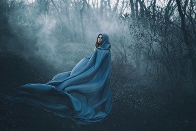 a big, beautiful woman in a blue raincoat, walks in a fog. background dark forest, bare trees. creative colors