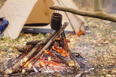 tourist kettle on camp fire with tent on background. hand with stick about fire.