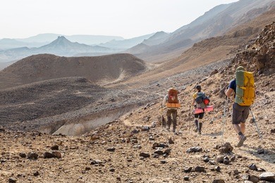 three backpackers tourists group walking hiking rocks desert path trail marking sign purple trailblazing marker, , ramon crater valley, israel tourism traveling.