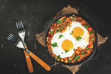 traditional mexican dish huevos rancheros - scrambled eggs with tomato salsa, with taco tortillas, fresh vegetables and parsley. breakfast. on a black concrete table. with a forks, copy space
