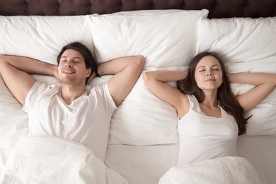 young happy married couple wearing white resting with eyes closed in bed, lying with hands behind the head after waking up in the morning, or going to sleep at night. lovers relaxing in bed. top view
