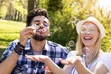 couple sitting on the park bench and blowing soup bubbles on beautiful sunny day