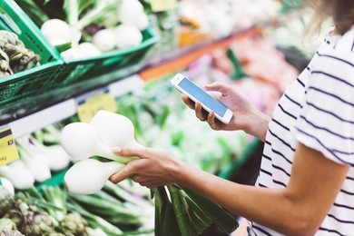 young woman shopping healthy food in supermarket blur background. female hands buy onion products and using smart phone in store. hipster at grocery holding basket. person comparing price of produce