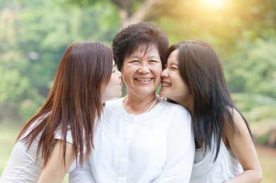portrait of beautiful asian daughters kissing elderly mother, senior adult woman and grown child. outdoors family at nature park with beautiful sun flare.