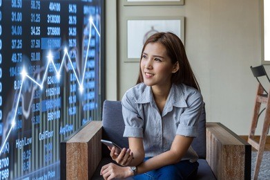 happy asian businesswoman sitting on working space and looking the trading graph over the stock exchange trading graph background in the modern office or coffee shop, business financial concept
