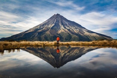 a mountaineer with an orange dress in a symmetric picture looking to the taranaki volcano in the north island of new zealand and with the reflection of tho mountain and the climber in a lake. zeland