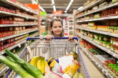 sale, consumerism and people concept - happy little girl with food in shopping cart at grocery store