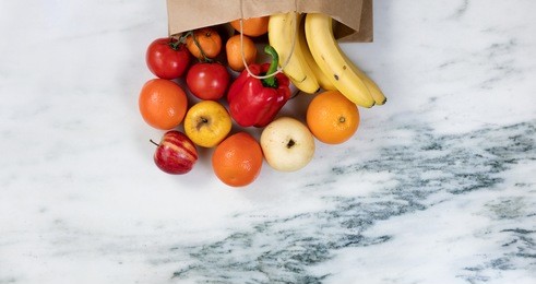flat lay view of fruits and vegetables spilling out of a paper bag onto marble stone countertop