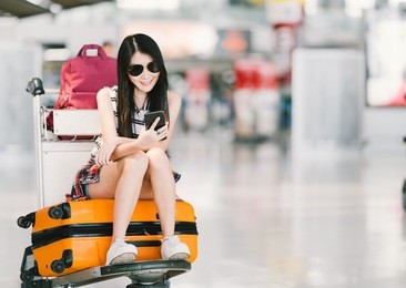 young asian girl using smartphone, waiting for flight at airport, sitting on baggage trolley or luggage cart. mobile communication technology or travel abroad concept, with copy space