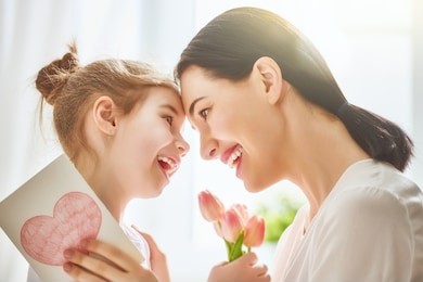 happy mother's day! child daughter congratulates mom and gives her flowers tulips and postcard. mum and girl smiling and hugging. family holiday and togetherness.