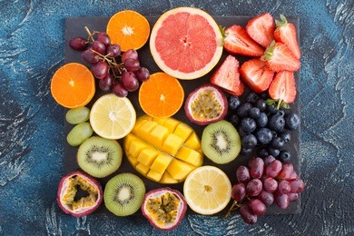 raw fruits platter: oranges, lemons, kiwis, grapefruits, strawberries, grapes, blueberries, passion fruit, mango on the black slate on the dark blue background, top view