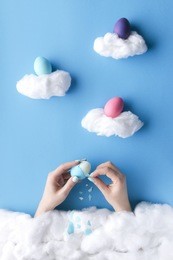 two beautiful women hands coming out the cotton wool clouds, peeling a blue easter egg on a blue sky paper background, in a heavenly scenery.