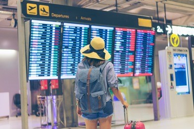 young woman traveler in international airport looking at the flight information board holding suitcase or baggage in her hand, checking her flight at the airport terminal. 