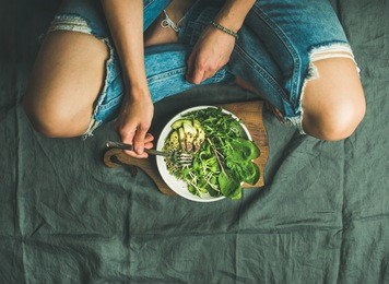 green vegan breakfast meal in bowl with spinach, arugula, avocado, seeds and sprouts. girl in jeans holding fork with knees and hands visible, top view, copy space. clean eating, detox food concept