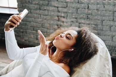 happy modern african american young girl talking on video chat and lying at home with loft interior, communication concept. smiling hipster girl making self portrait on her telephone at home