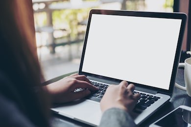 mockup image of business woman using and typing on laptop with blank white screen and coffee cup on glass table in modern loft cafe
