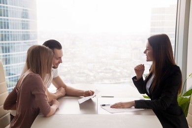 side view of realtor and young couple sitting at office desk discussing property for sale. potential buyers holding house plan, considering investment in home or taking loan to purchase real estate 
