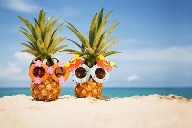 couple of funny attractive pineapples in children's cheerful sunglasses on sand against turquoise sea. tropical summer vacation concept. happy sunny day on the beach of tropical island. 