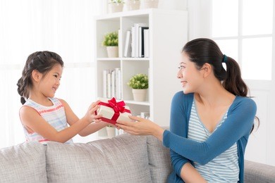 mixed race chinese smiling daughter giving her mother a red ribbon gift box want to give mom a surprise at mother's day at home holiday in the afternoon.