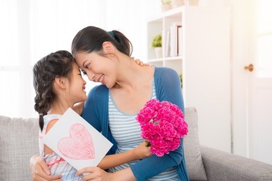 young asian mother accepting the little daughter hand painted card, embracing, little girl holding a pink flower girl, sitting on the sofa in the living room, happy looking at each other, copy space for mother's day.