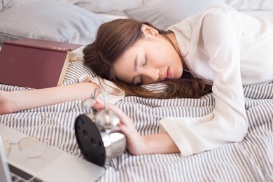 beautiful and attractive asian woman lying down on the bed while she sleeping with her notebook or laptop and her hand touching the vintage alarm clock feel not want to wake up at the morning