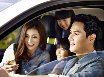 happy asian family with two children traveling by car, mother using cellphone while father driving, focus on the father.