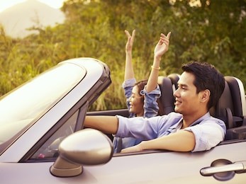young asian couple riding in a convertible sport car enjoying the cool breeze at sunset.