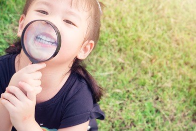 little girl child holding magnifying glass and showing front teeth with big smile on green grass: healthy happy funny smiling face young adorable lovely female kid with new tooth dental loss.