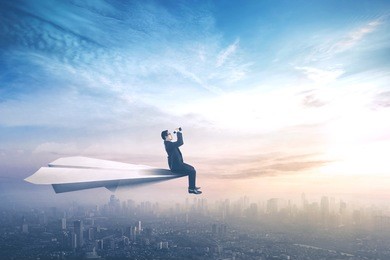 young businessman sitting on a paper aircraft while flying above a city and looking through binoculars