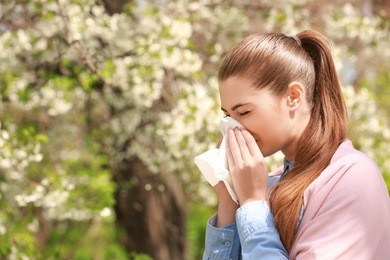 sneezing young girl with nose wiper among blooming trees in park