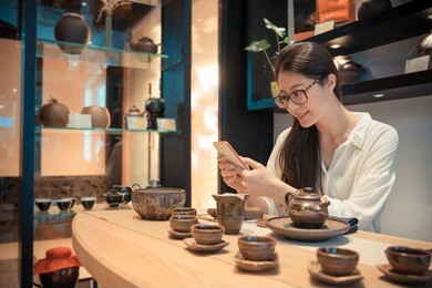 smiling woman using her mobile cell phone to take pictures record traditional chinese tea culture when sitting in ceramic tea group table at home.