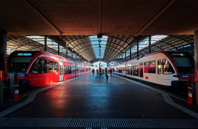 perspective view of a platform in lucerne central railway station with sunlight cast on trains parking by the platform & passengers hurrying for boarding~a beautiful corner in lucern railroad terminal