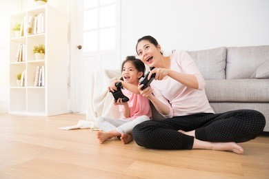 good relationship cute little girl with young mother using joystick playing video game sitting together in living room wooden floor enjoying family holiday.