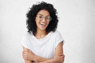 cheerful young good looking woman with clean dark skin and black shaggy hair posing indoors with crossed arms, smiling broadly with her white straight teeth, laughing at good joke, wearing casual top