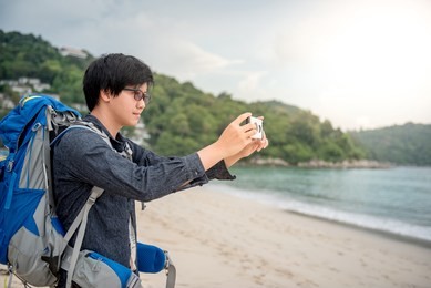 young asian backpacker man take photos of beach and sea by smartphone, summer holiday vacation and travel tropical island concepts