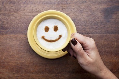 hand holding a cute yellow cup with coffee cream. food art creative concept image, happy face drawing with cinnamon powder over wooden background.