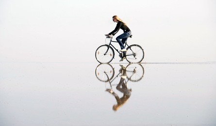 a girl riding a bicycle by water, that creates a bewitching mirror illusion.