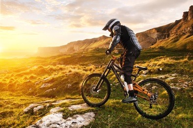 the rider in full protective equipment on the mtb bike is riding toward the sunset in the rays of the sunset sun against the background of the rocks of the setting sun and clouds
