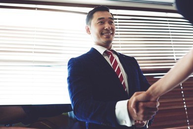 portrait of a smiling young managing director shaking hands with his business partners while standing in office space interior, asian male entrepreneur enters into an agreement with his new partner