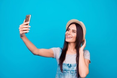 young cheerful attractive brunette  is smiling on the blue background. she is taking selfie on the camera of her phone, wearing casual summer outfit and a hat