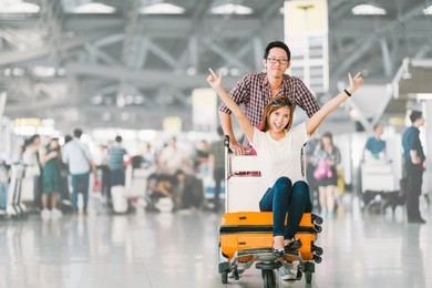 young asian tourist couple happy and excited together for the trip, girlfriend sitting and cheering on baggage trolley or luggage cart. holiday vacation traveling abroad concept, with copy space
