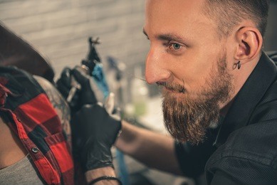 focus on side view portrait of man expressing thoughtfulness while doing tattoo on hand of woman. muse concept