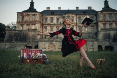 woman sorceress flies in air and reads book against backdrop of ancient castle. levitation. on grass there is suitcase with flowers.